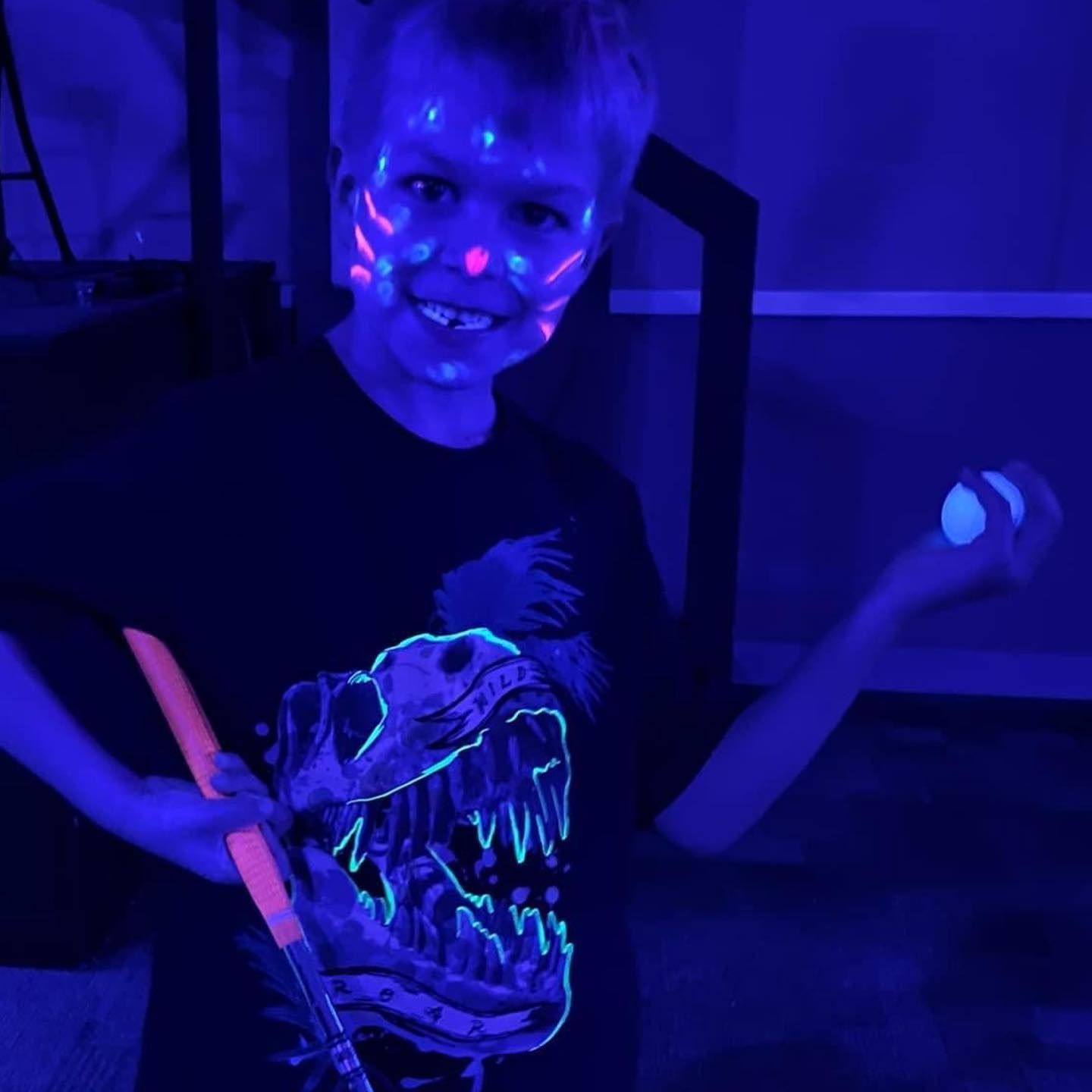 Boy with glowing face paint and dinosaur shirt under black light, holding paint brush and glowing objects.