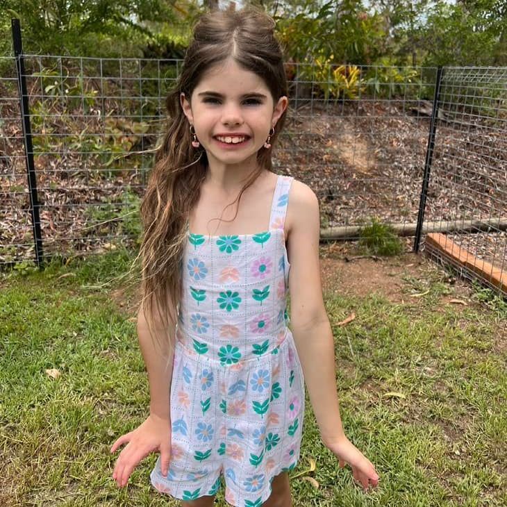 Girl in white romper with floral pattern, smiling outdoors. Green grass, fence background.