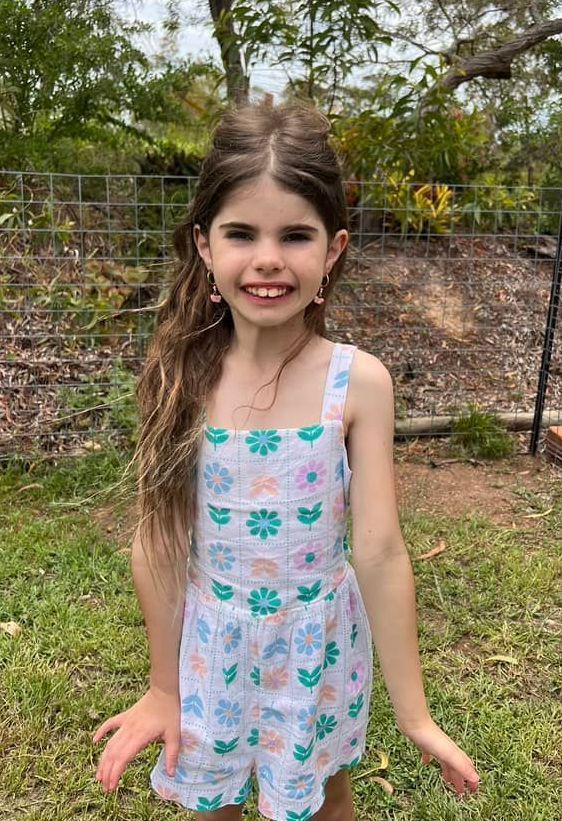 Girl in a floral romper smiles outdoors, arms out, against a fence and greenery.