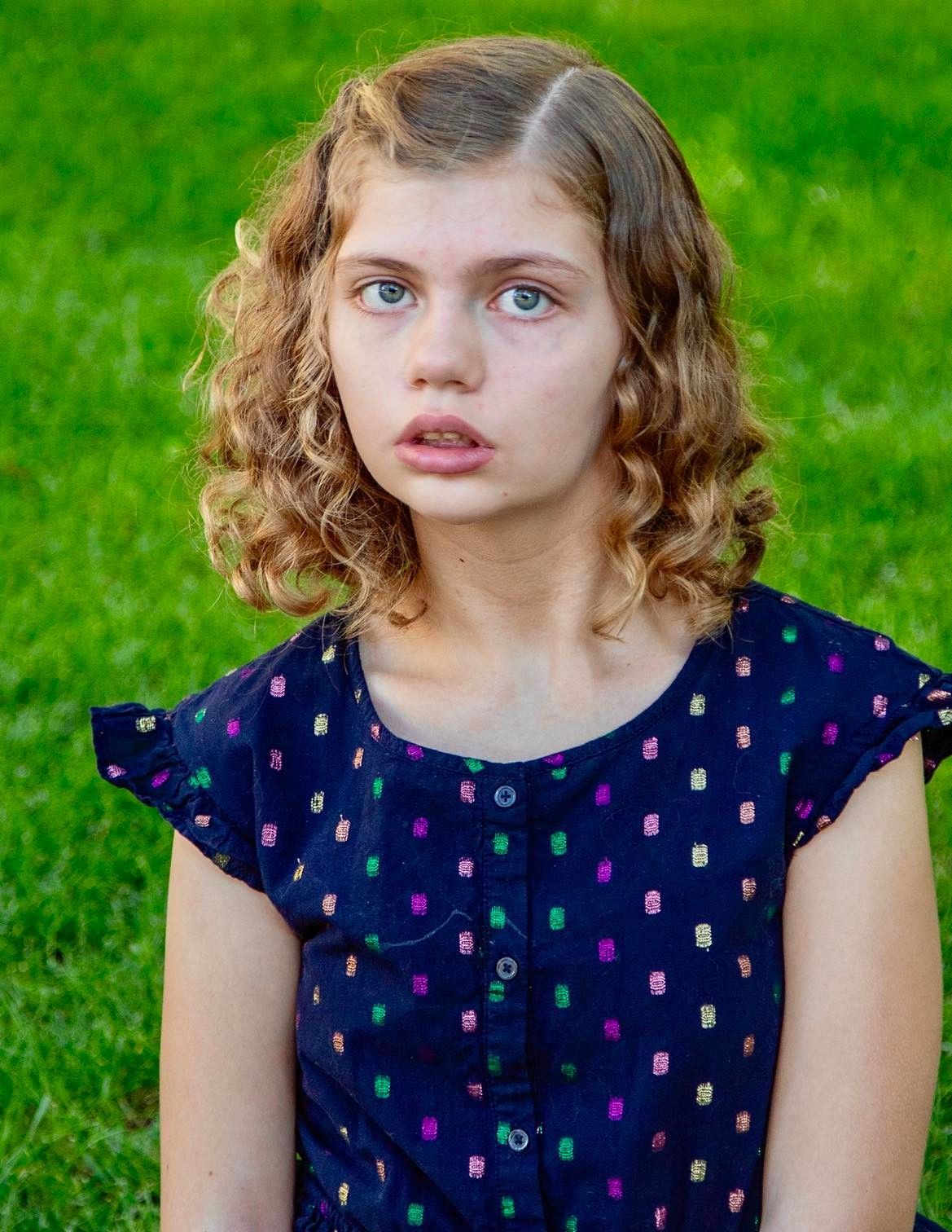 Girl with curly brown hair wearing a blue patterned dress, looking directly at the viewer outdoors.