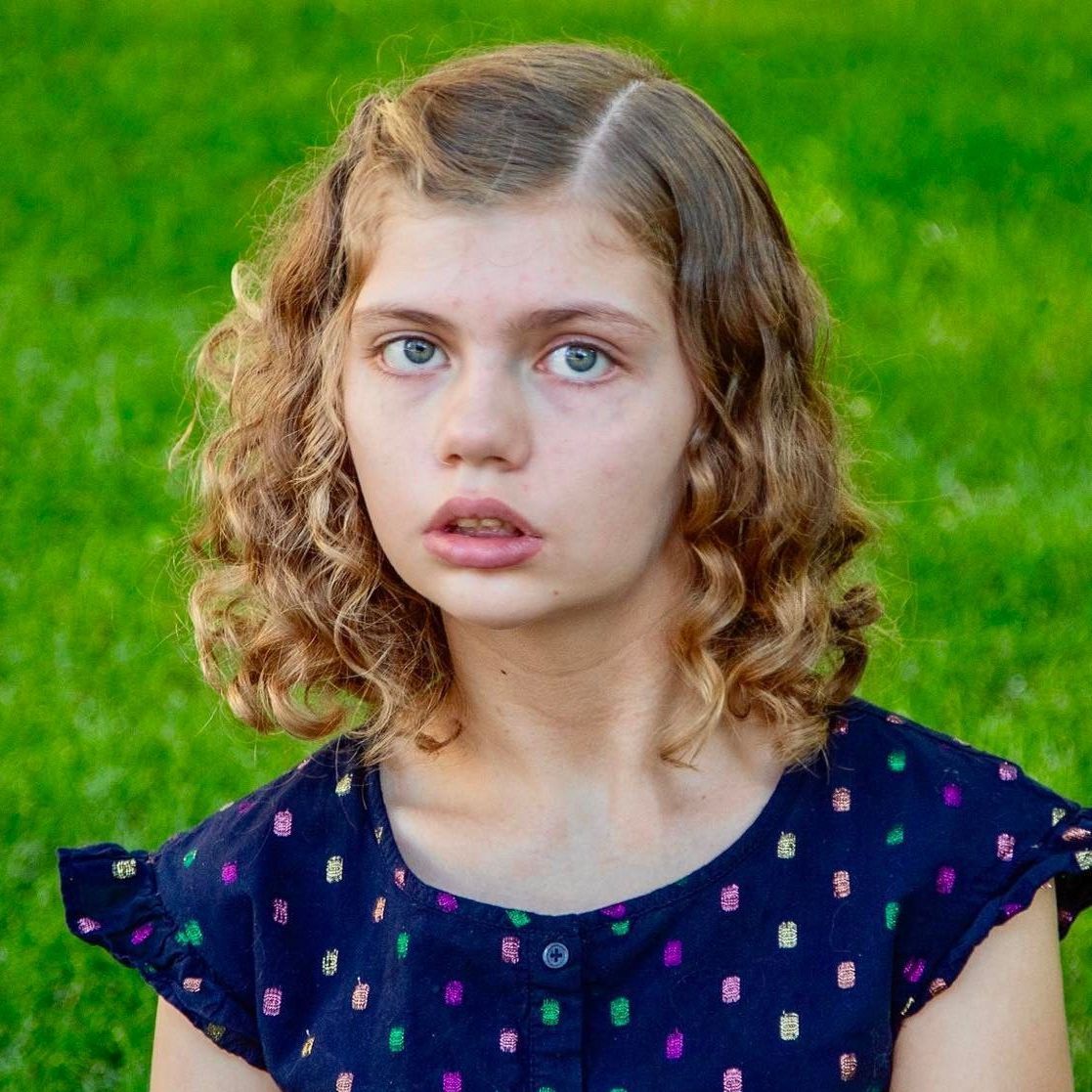 Girl with curly hair wearing a blue patterned shirt, looking directly at the viewer with a neutral expression.