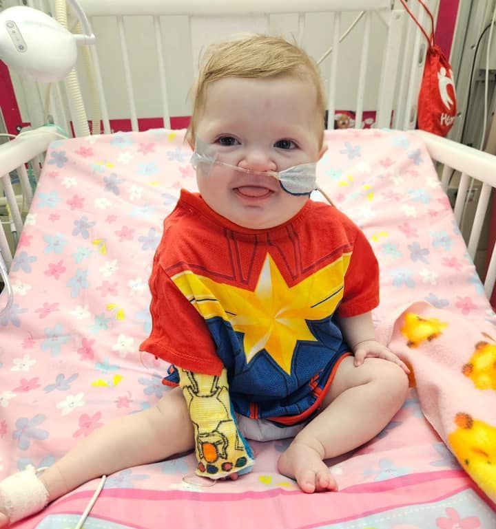 Child in Captain Marvel shirt with medical tubing in a hospital crib, smiling with tongue out.