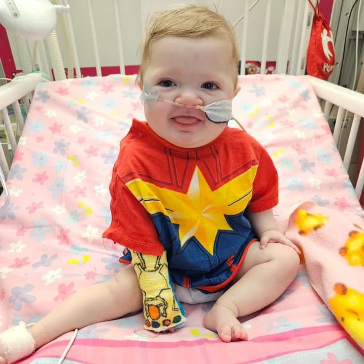 Baby in a Captain Marvel shirt sits in a hospital crib with medical tubing.