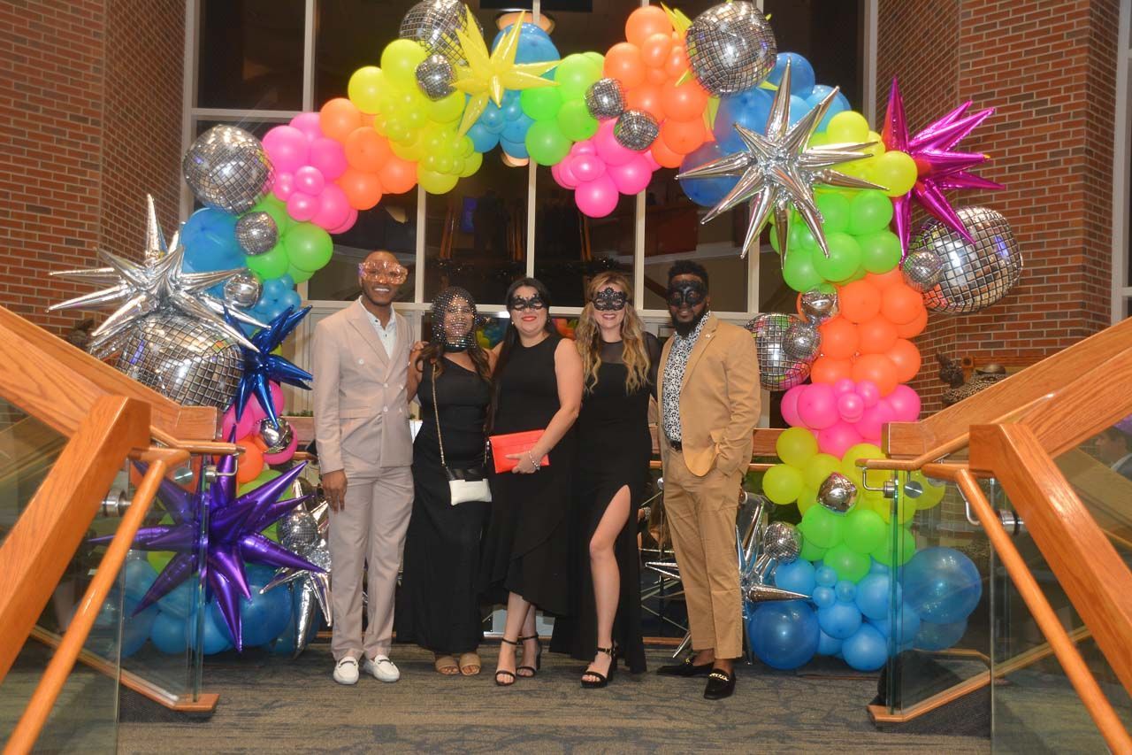 Five people pose beneath a colorful balloon arch. Two wear masks.