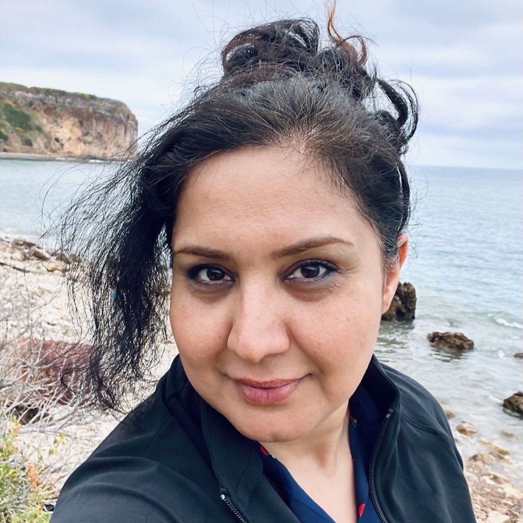 Woman with dark hair looking at the camera near a rocky shore with the ocean and a cliff in the background.
