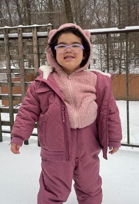 Child in pink snowsuit and cat-ear hood smiles outside in snowy setting.