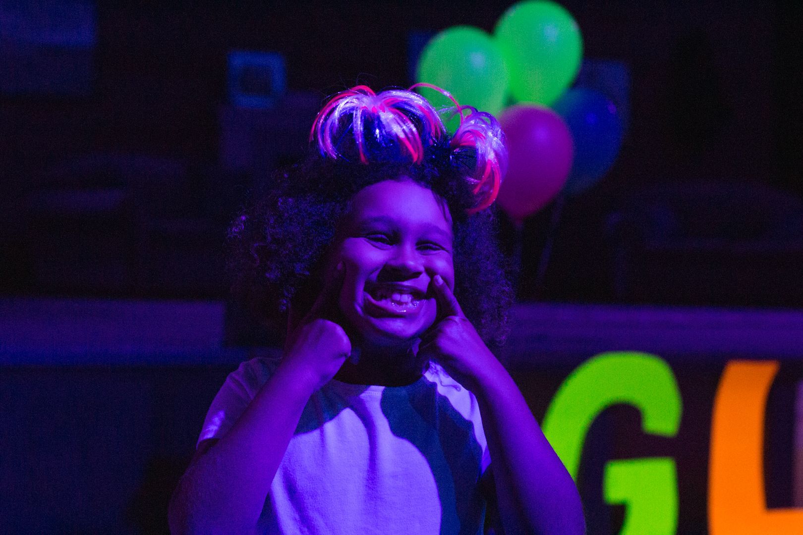 Girl smiling, holding cheeks under black light, glowing headband, balloons.