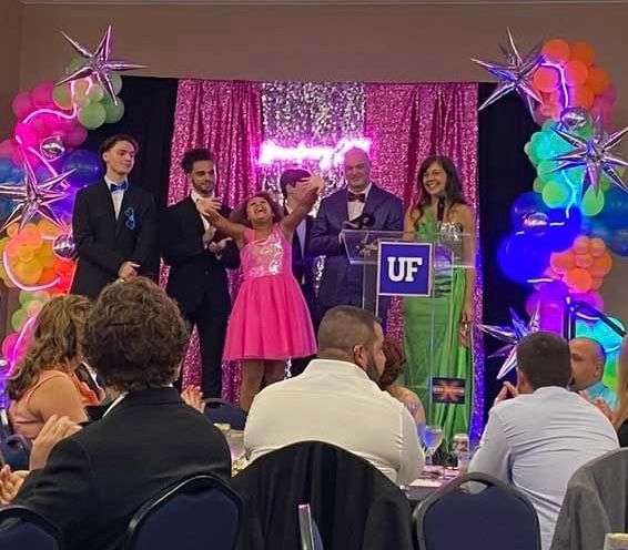 People on stage at event with pink sequins, neon sign, star balloons, and crowd.