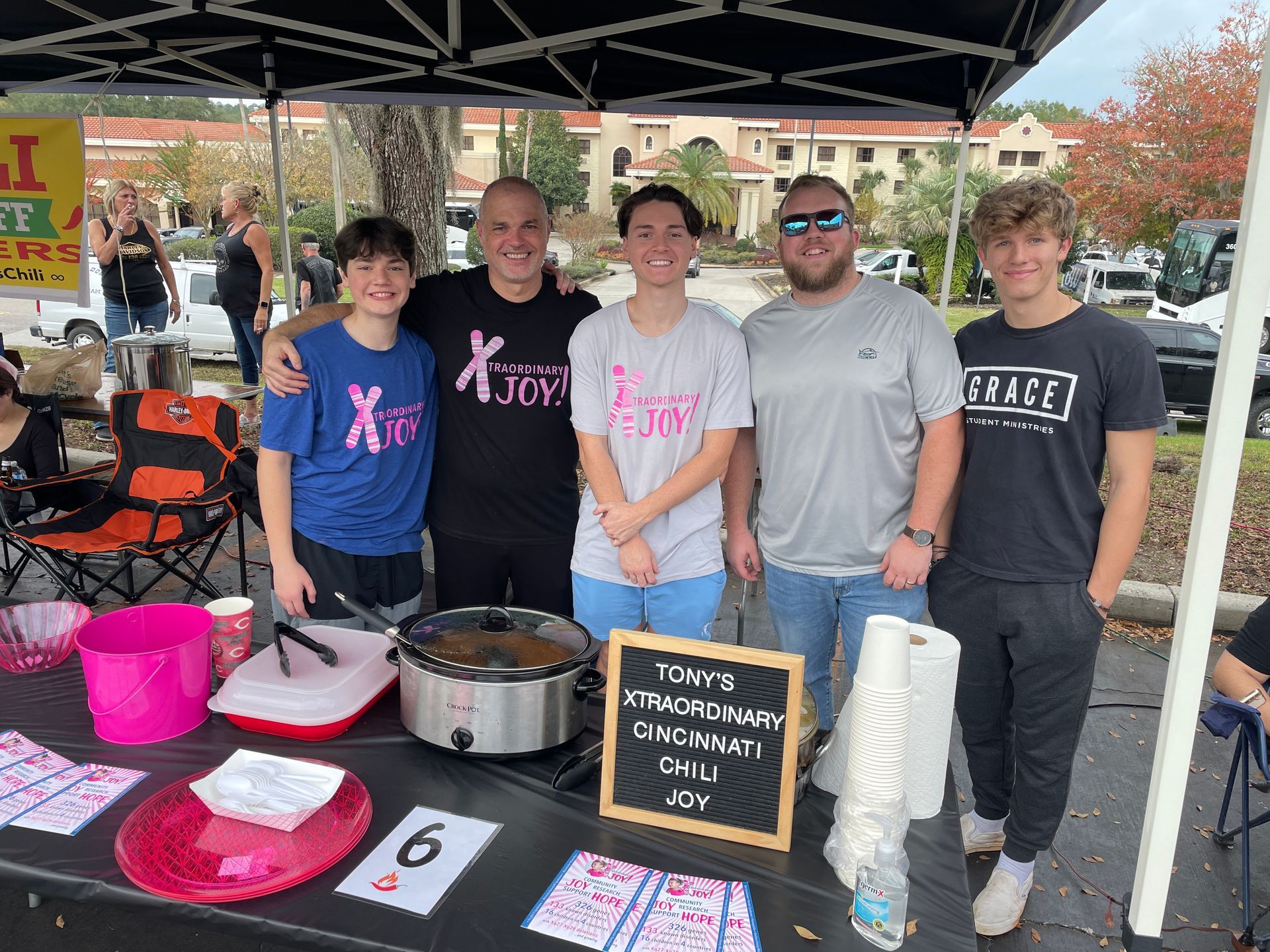 People at a chili fundraiser. Table displays chili, pink buckets, and sign reading 