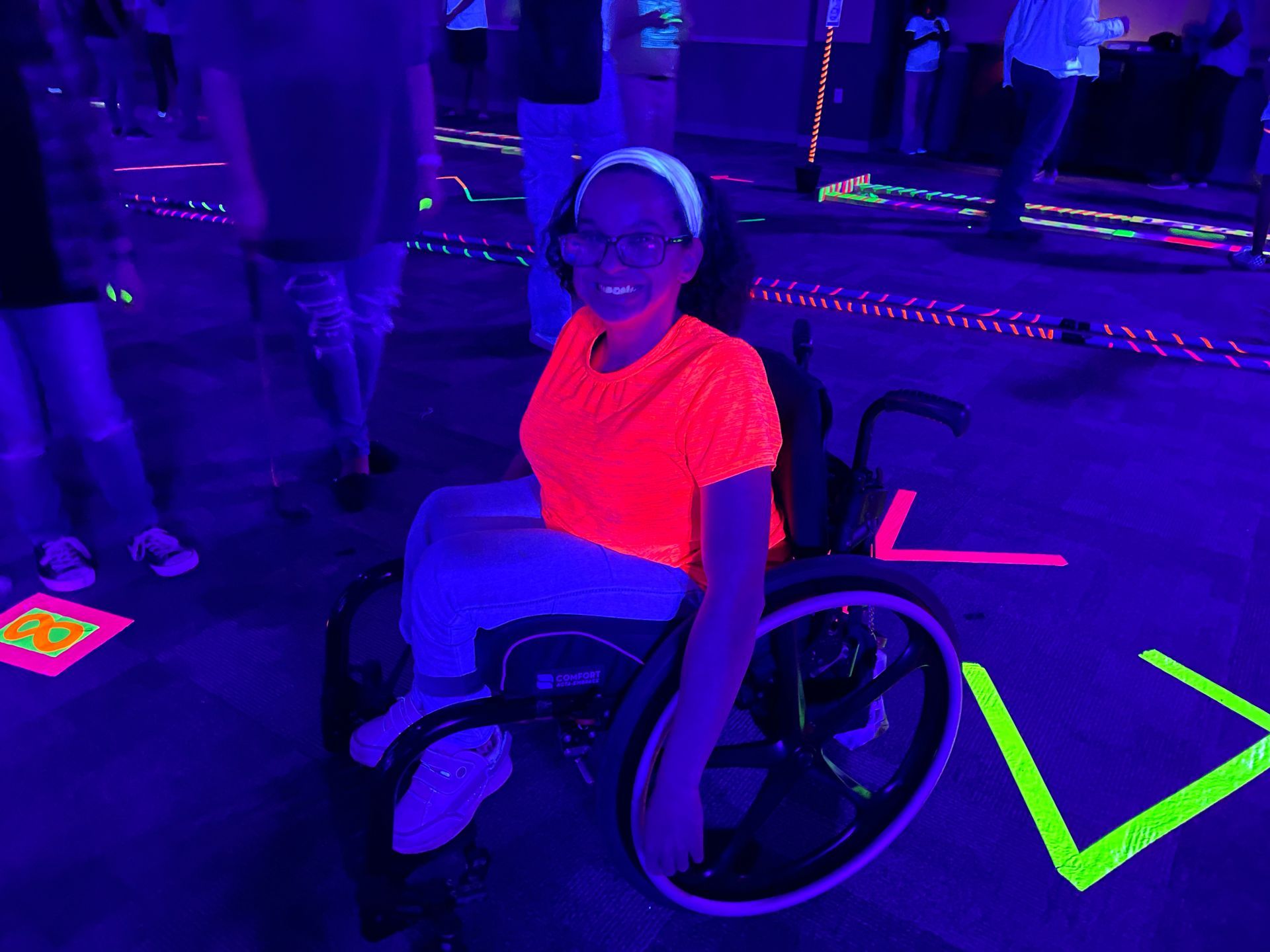Person in wheelchair at a blacklight event, wearing neon orange top and white headband.