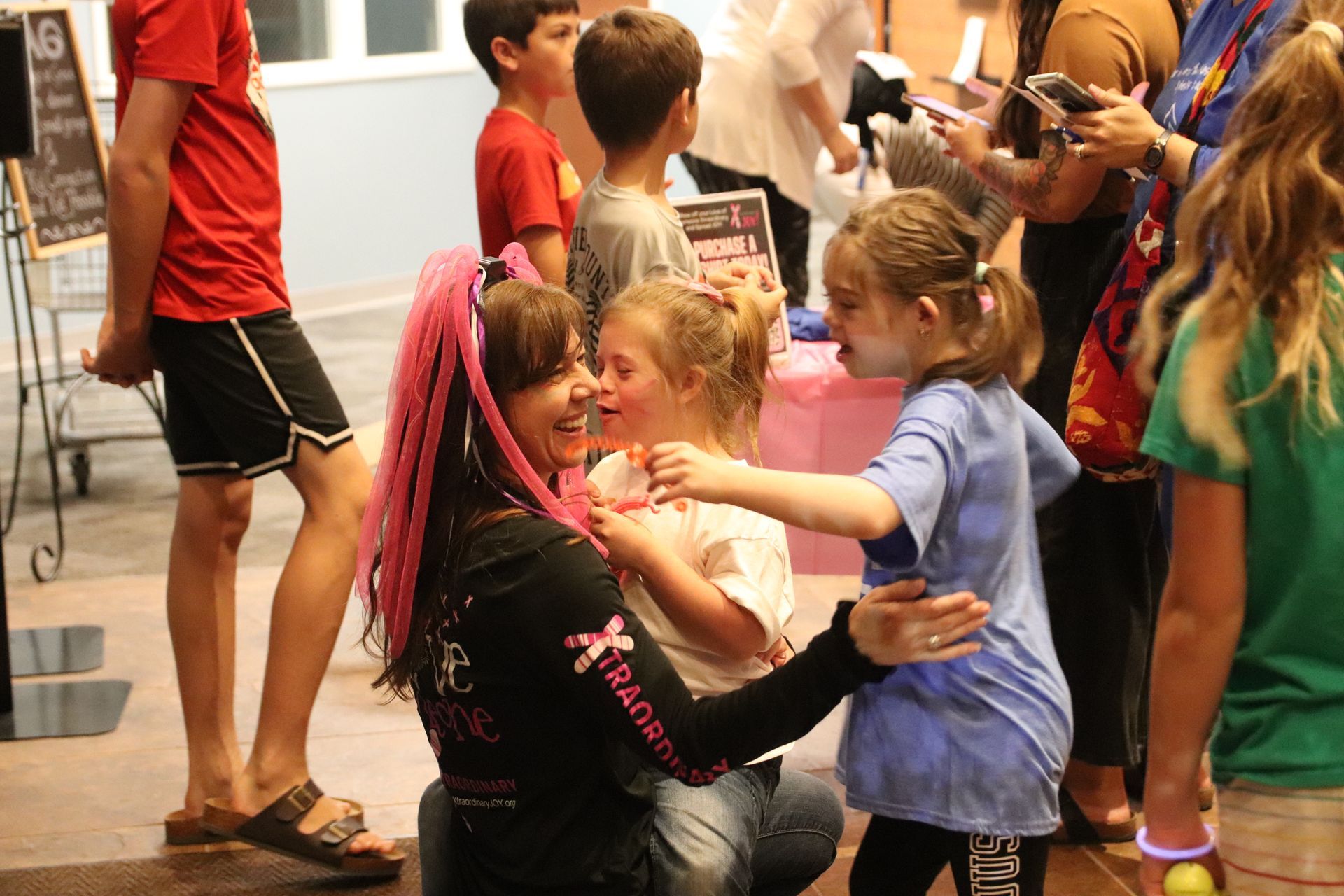 Woman with pink hair hugs two children at an event, surrounded by other people.