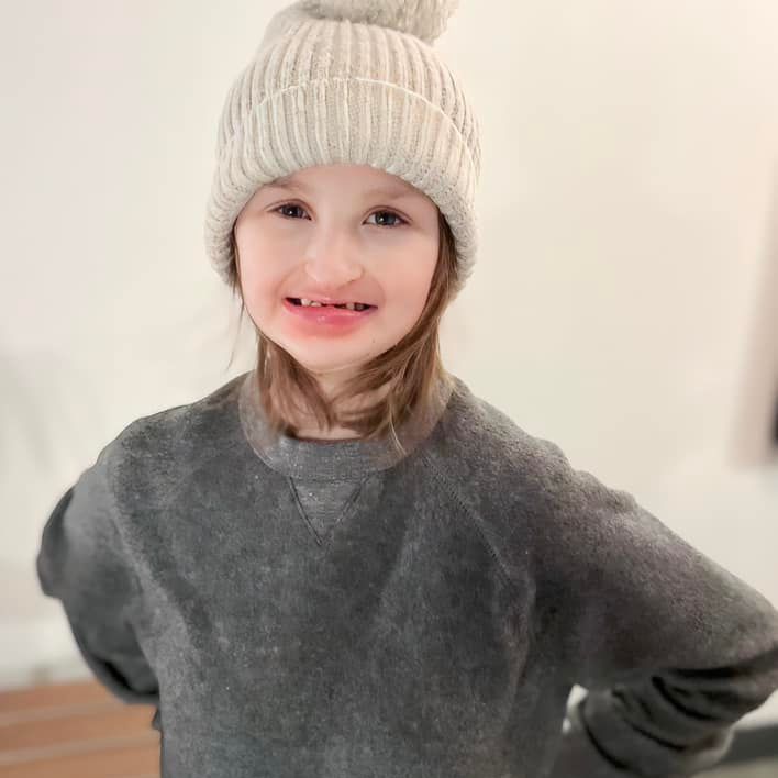 A child wearing a gray sweater and cream-colored beanie smiles, arms on hips, against a white wall.