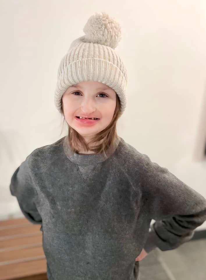 Girl wearing a gray sweater and a light-colored pom-pom beanie, smiling with hands on hips.