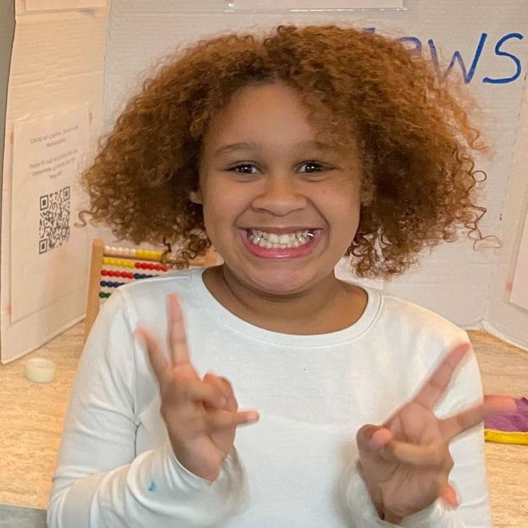 Girl with curly red hair smiles and holds up her hands in a peace sign pose.