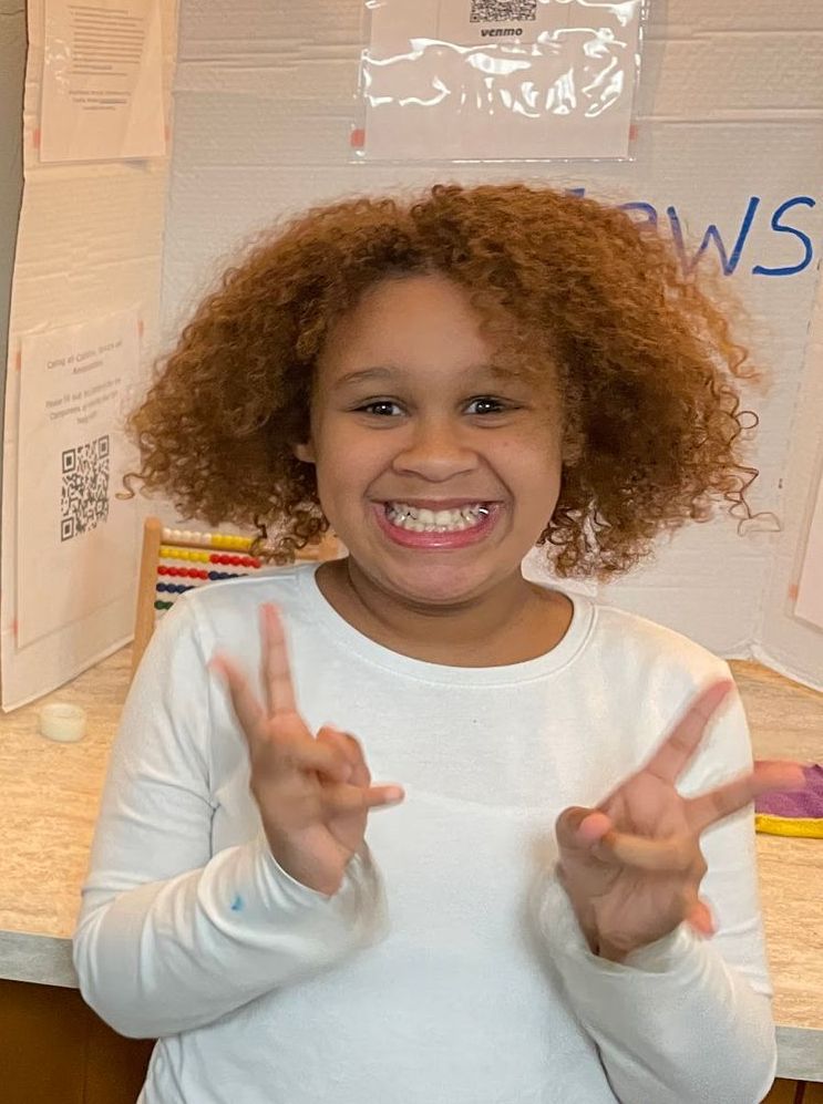 Young person with curly hair smiling, holding up two fingers on each hand. Background has a poster.