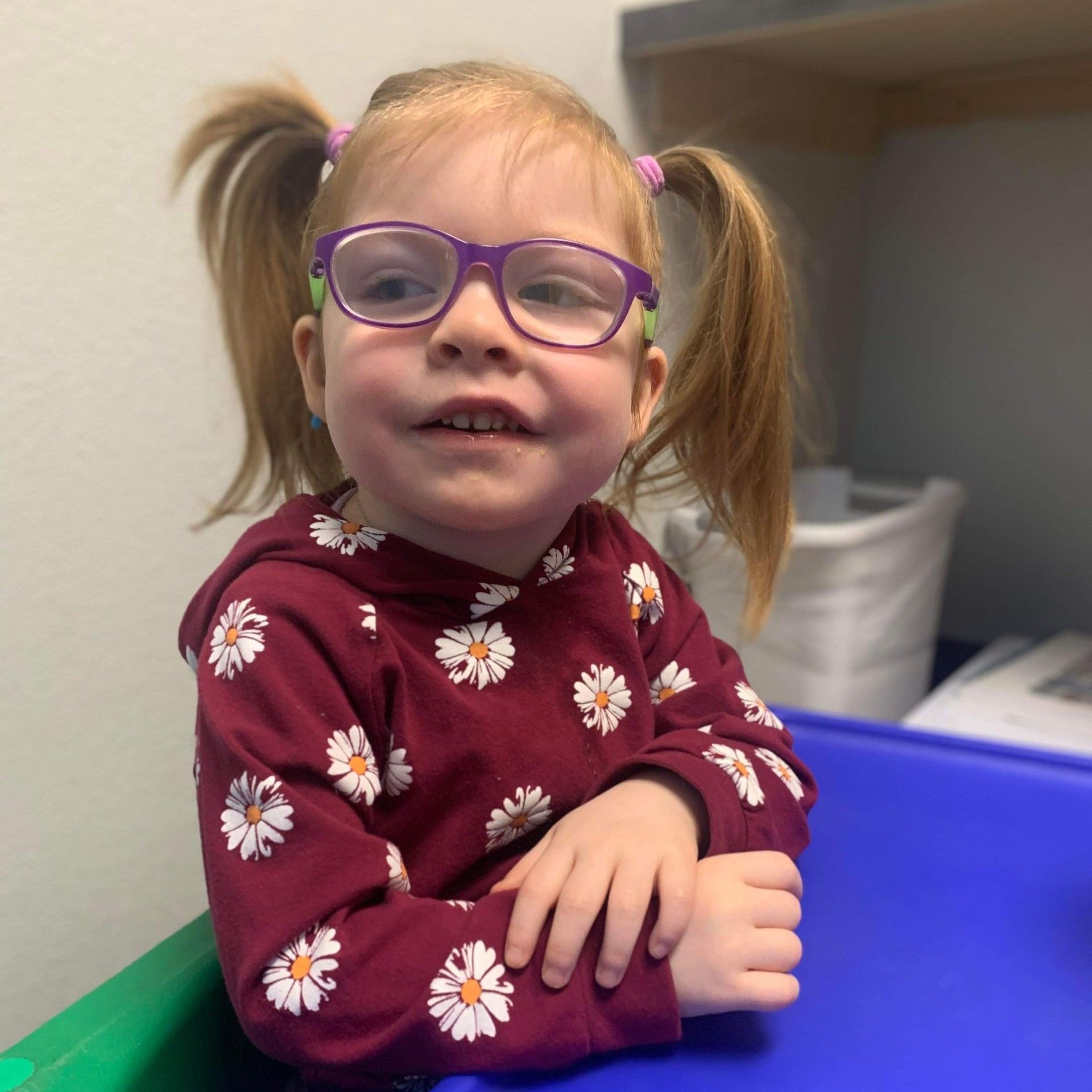 Young child with pigtails wearing glasses and a daisy-print hoodie, sitting at a blue table.