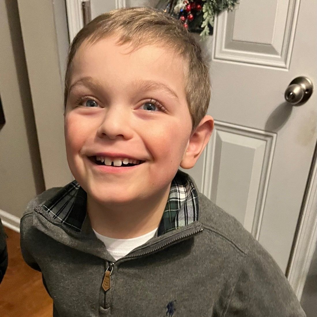 Boy with a big smile, wearing a gray zip-up sweater, standing in front of a white door.