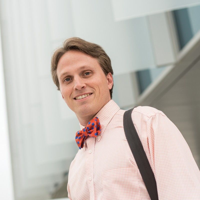 Man in light orange checkered shirt and patterned bow tie smiles, carrying a bag.