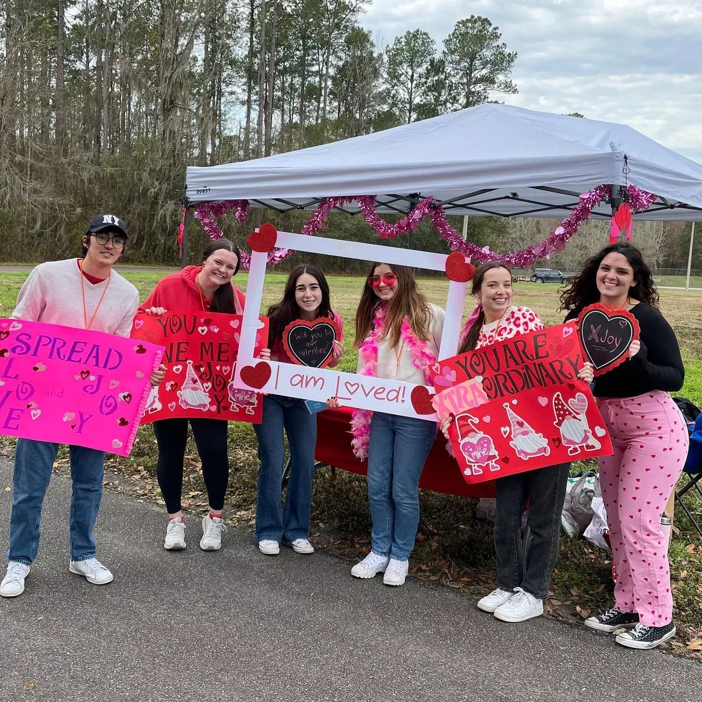 Group of people holding Valentine's Day signs and a photo frame near a decorated tent in a park.