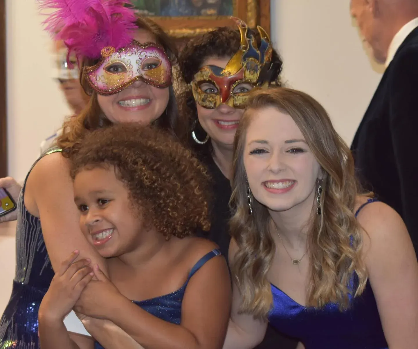 Four people at a party wearing masks, smiling. One child is hugging a woman in a blue dress.