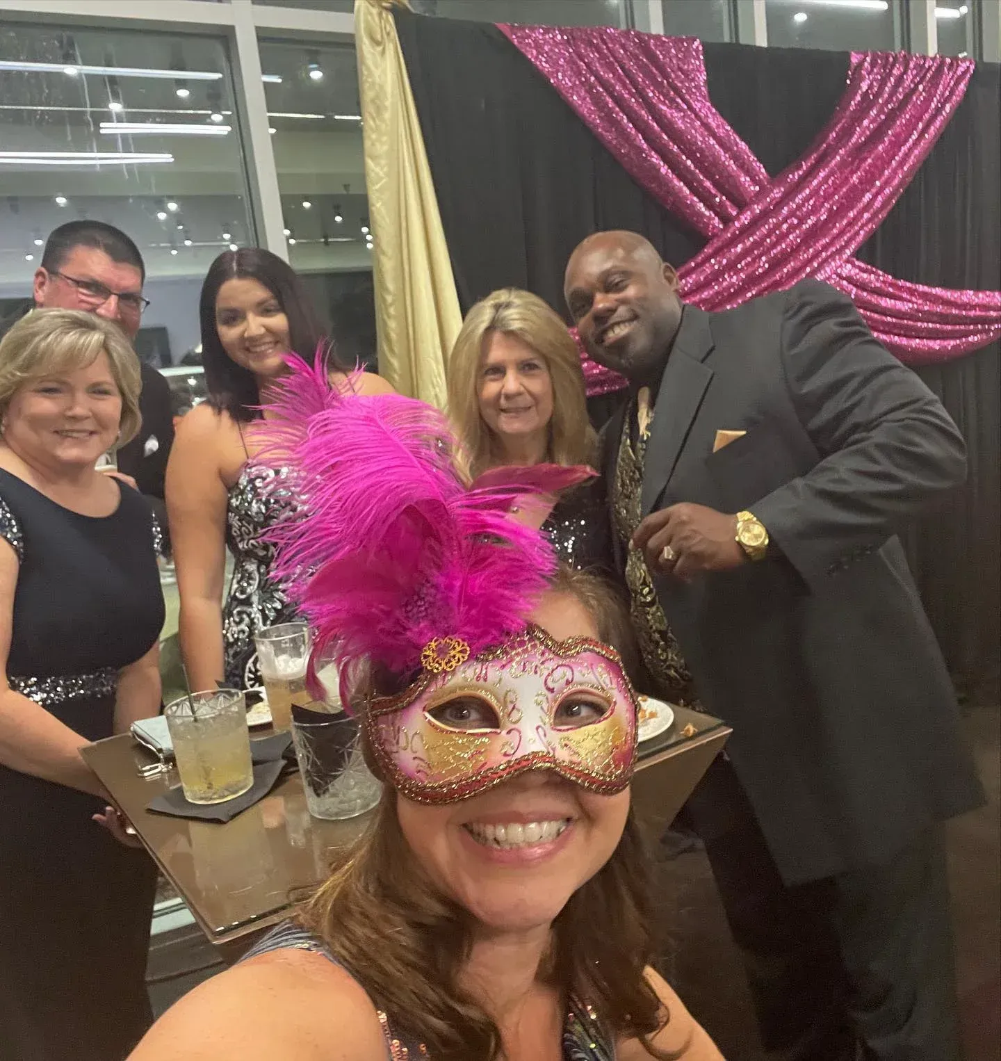 Group posing at a masquerade event; woman in foreground smiles, wearing mask with pink feathers, behind her, group poses.