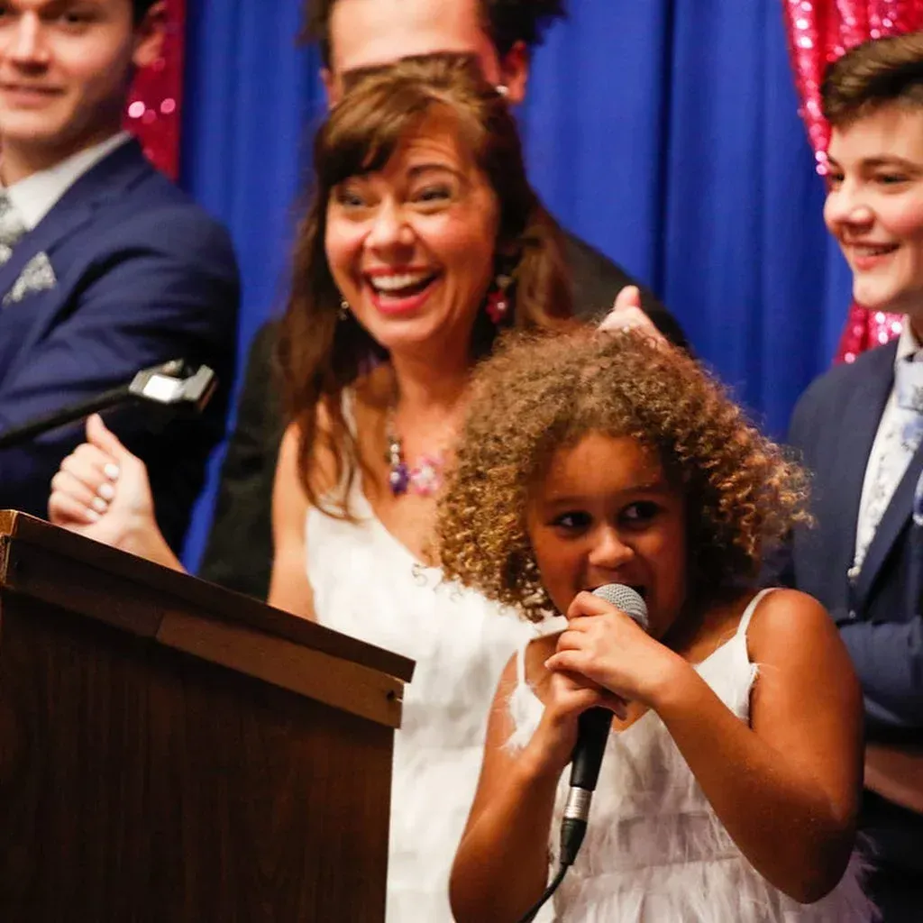 Young girl singing into a microphone at a podium, smiling. Adults in formal wear smile in the background.