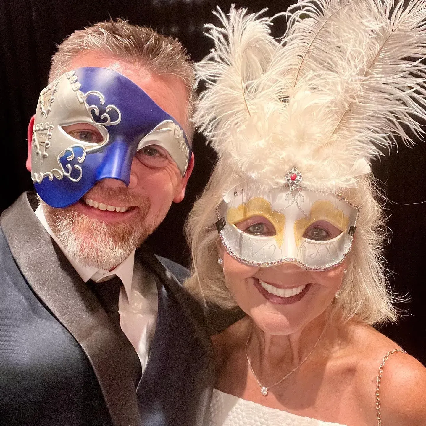 Man and woman in masquerade masks, smiling. Man in blue and silver mask, woman in white and gold mask with feathers.