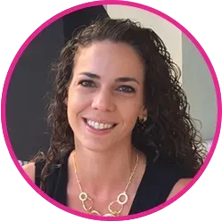 Woman with curly brown hair, smiling, wearing a black top and gold necklace, set in a room with a pink border.