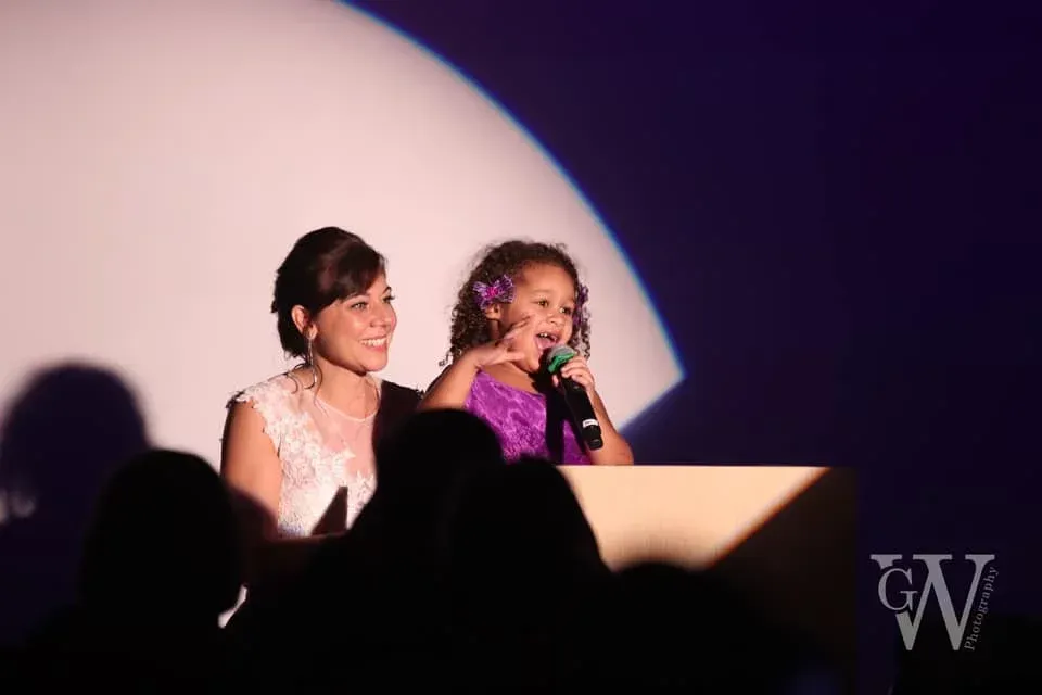Woman and girl at a podium under a spotlight; girl speaks into microphone, gesturing; audience in silhouette.