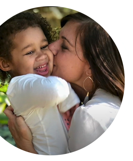 Woman kisses a laughing child outdoors. The child wears a white shirt.