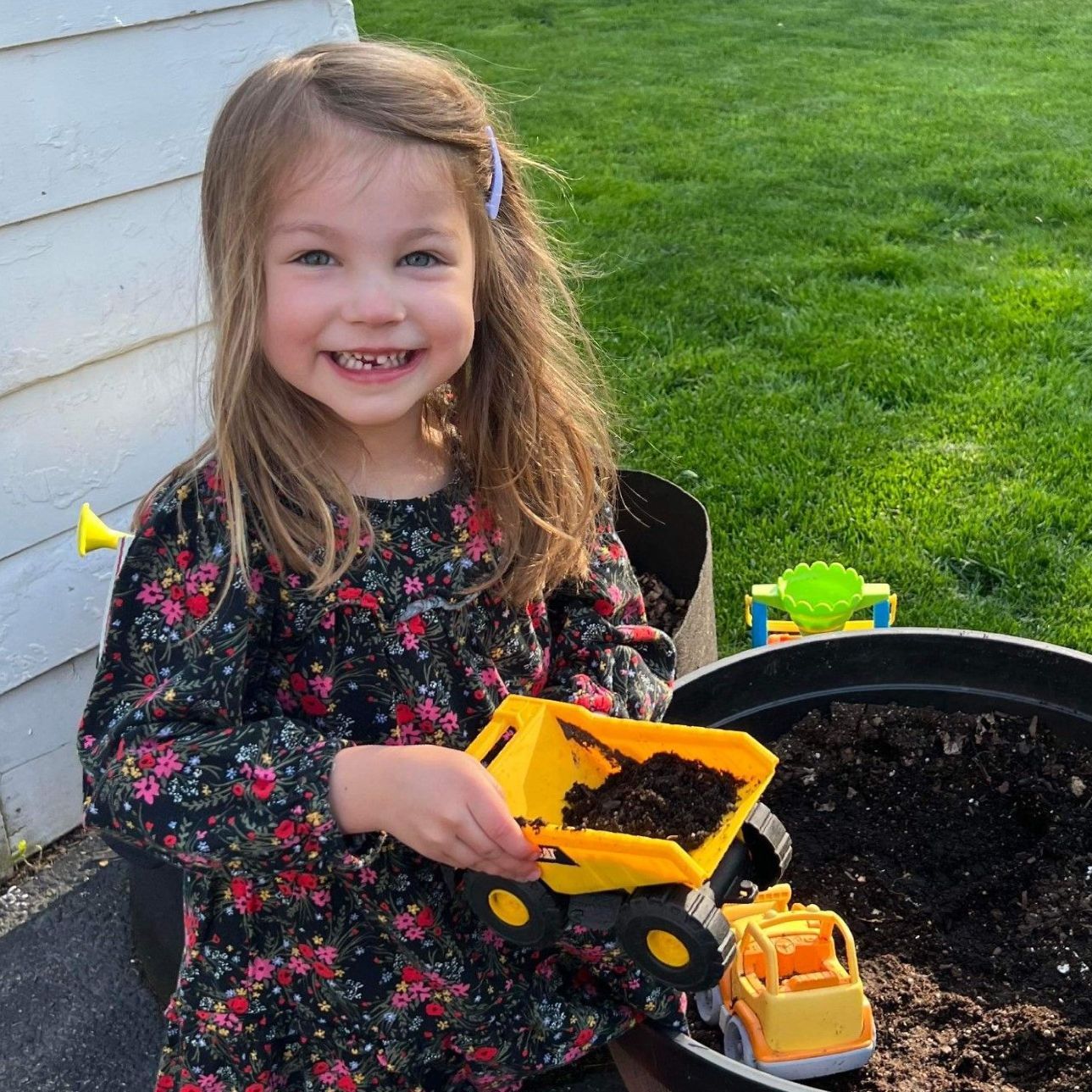 Girl smiling while holding a toy truck filled with dirt, next to a planter outdoors.