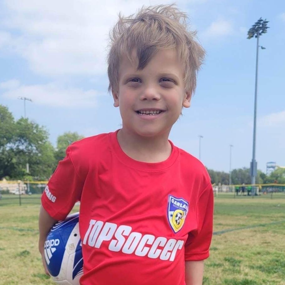 Boy in red soccer jersey holding ball, smiling outdoors on a field.