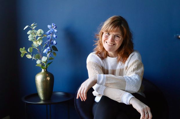 Vrouw met bril, glimlachend, zit bij een blauwe muur met een vaas bloemen.