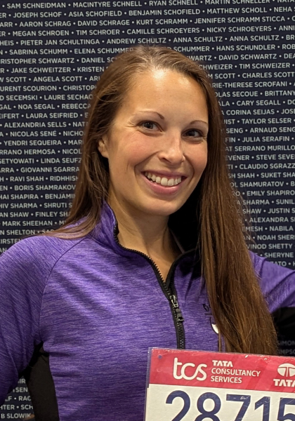 Smiling runner in purple jacket holding a red TCS race bib in front of a name wall