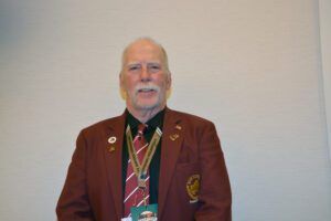 Man in a maroon blazer and patterned tie standing against a plain wall, smiling at the camera.