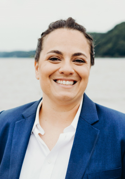 Smiling person in a blue blazer and white shirt outdoors by a body of water.