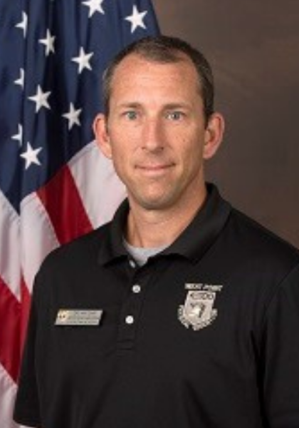 Man in black polo shirt standing before an American flag, facing the camera with a neutral expression