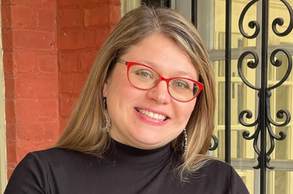 Smiling person with red glasses and a black top standing by a brick wall and wrought-iron gate