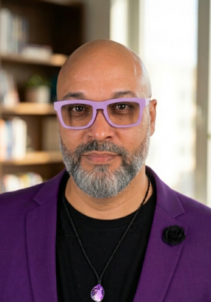 Man with purple glasses and blazer, wearing a pendant, against a softly blurred indoor background