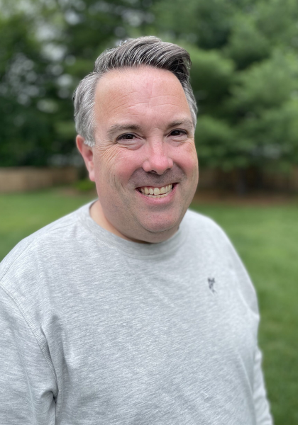 Smiling person in a light gray shirt standing outdoors with blurred green trees behind them