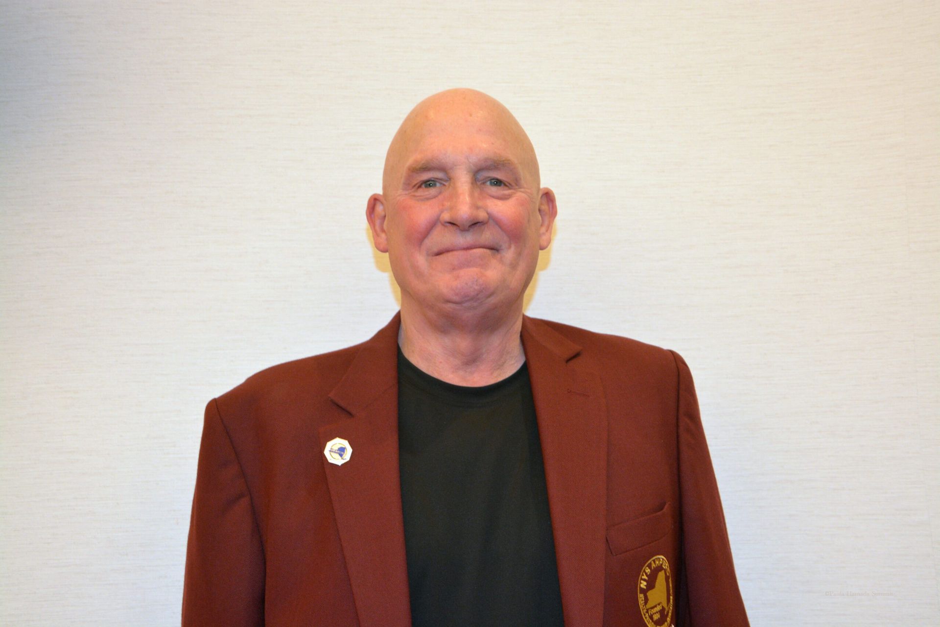 Smiling bald man in a maroon blazer standing against a plain wall.