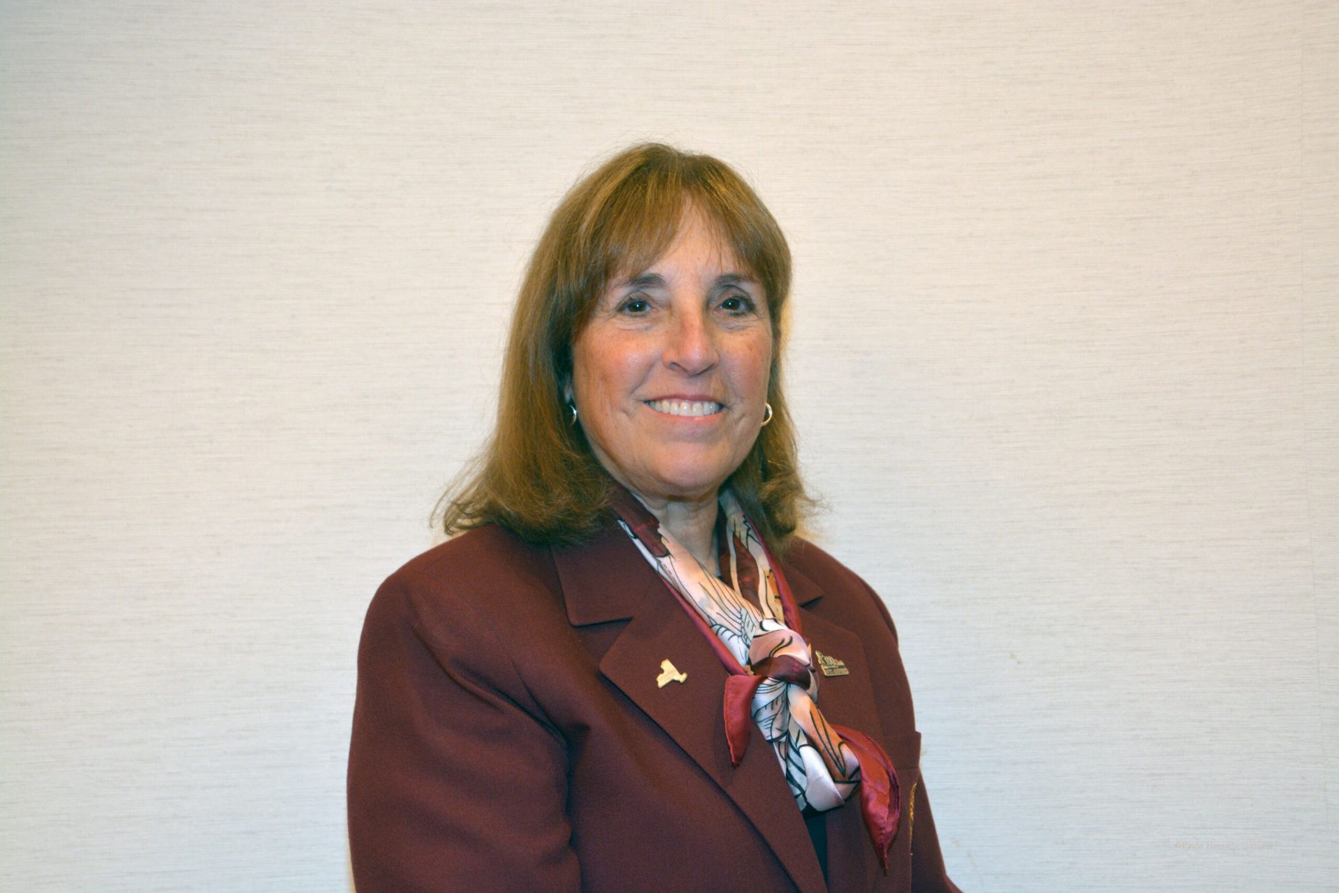 Woman in a maroon blazer and patterned scarf standing against a plain wall, smiling at the camera.