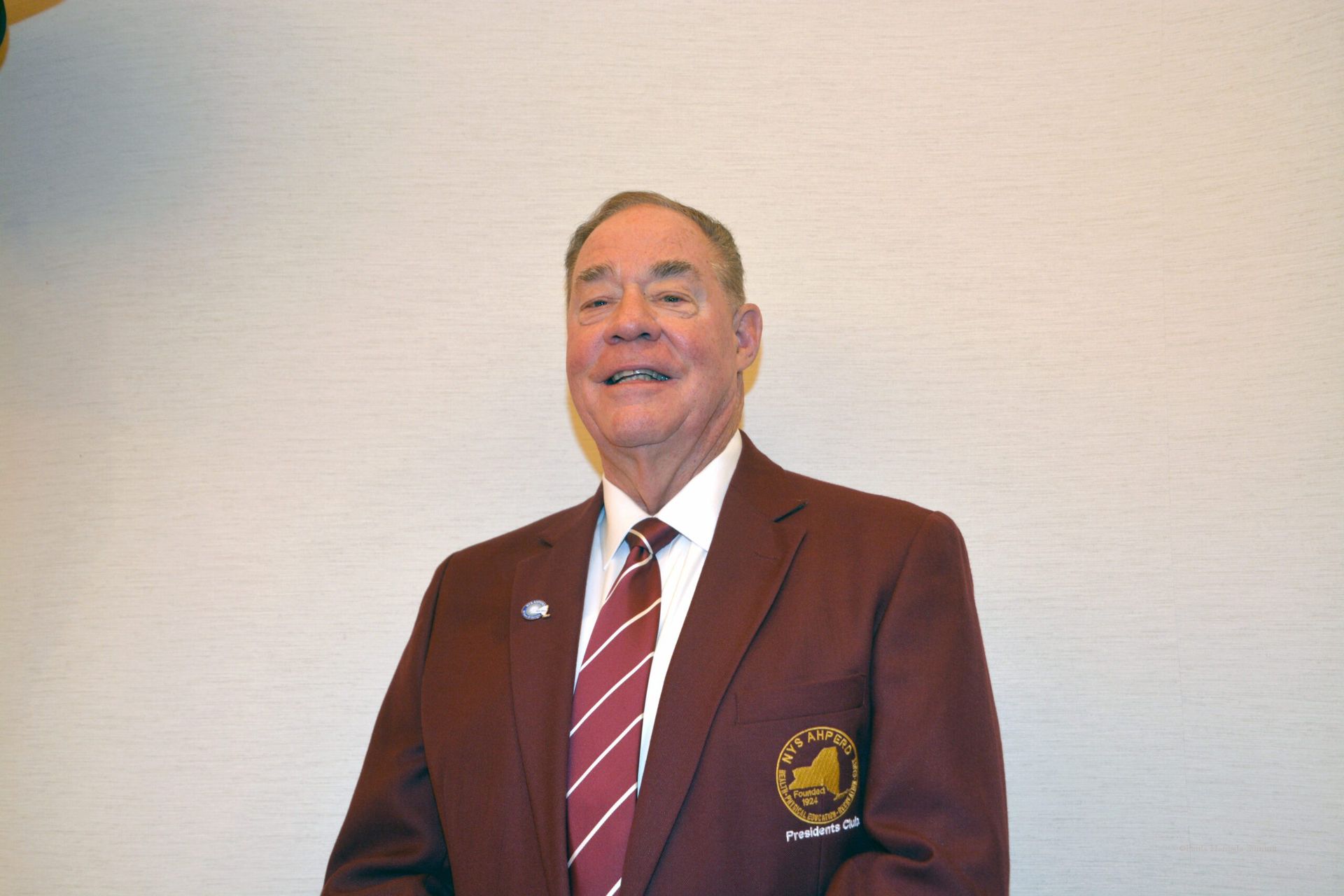 Man in a maroon blazer with a striped tie, smiling against a plain wall.