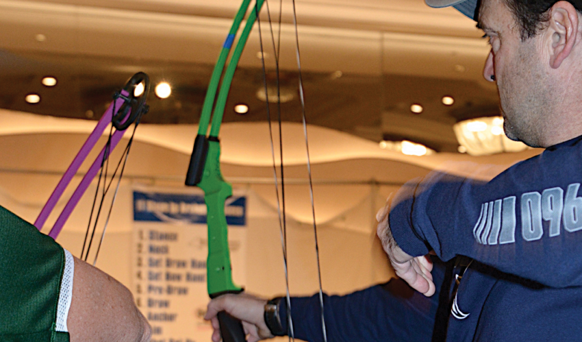Person drawing a green and purple bow in an indoor archery range