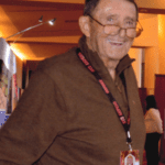 Smiling man in a brown shirt and lanyard standing indoors under warm orange lighting