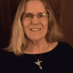 Smiling person with glasses and shoulder-length hair wearing a dark top and cross necklace.