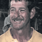 Smiling man in a light shirt at a dimly lit event, with a blurred crowd behind him