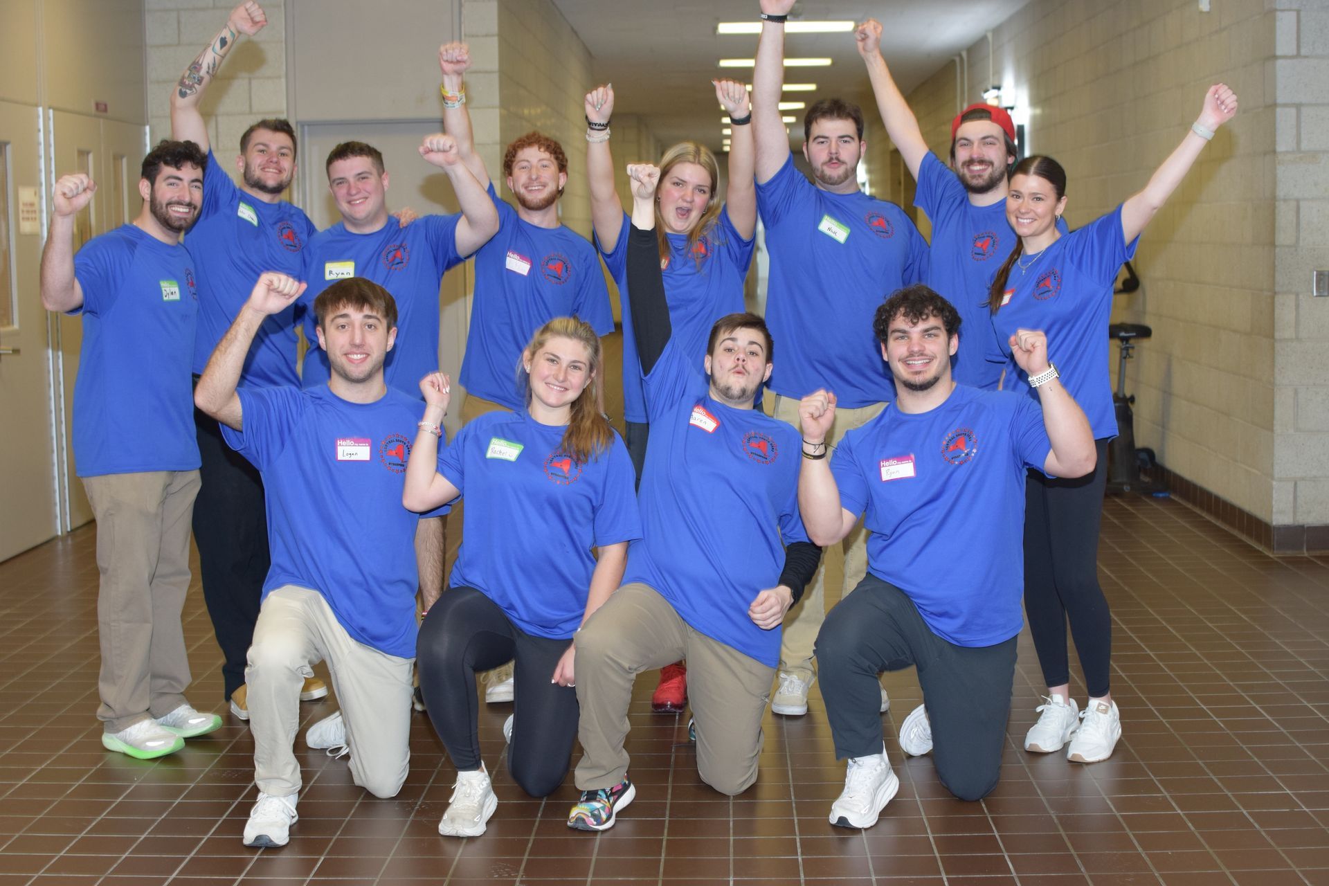Group of people in blue shirts posing indoors with raised fists in a hallway