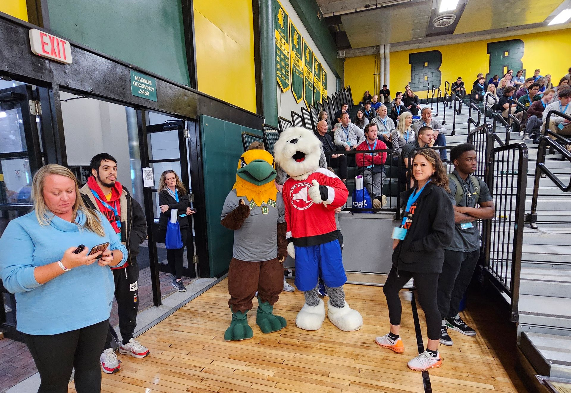 Mascots posing on a gym floor with bleachers full of spectators in the background