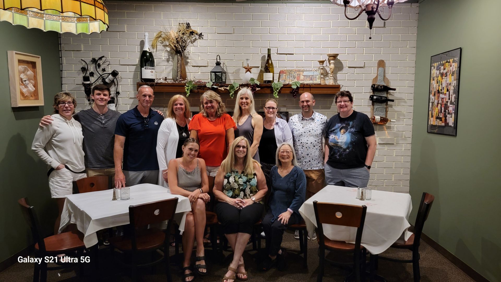 Group posed in a restaurant dining room, standing and seated around white-clothed tables, smiling at the camera.